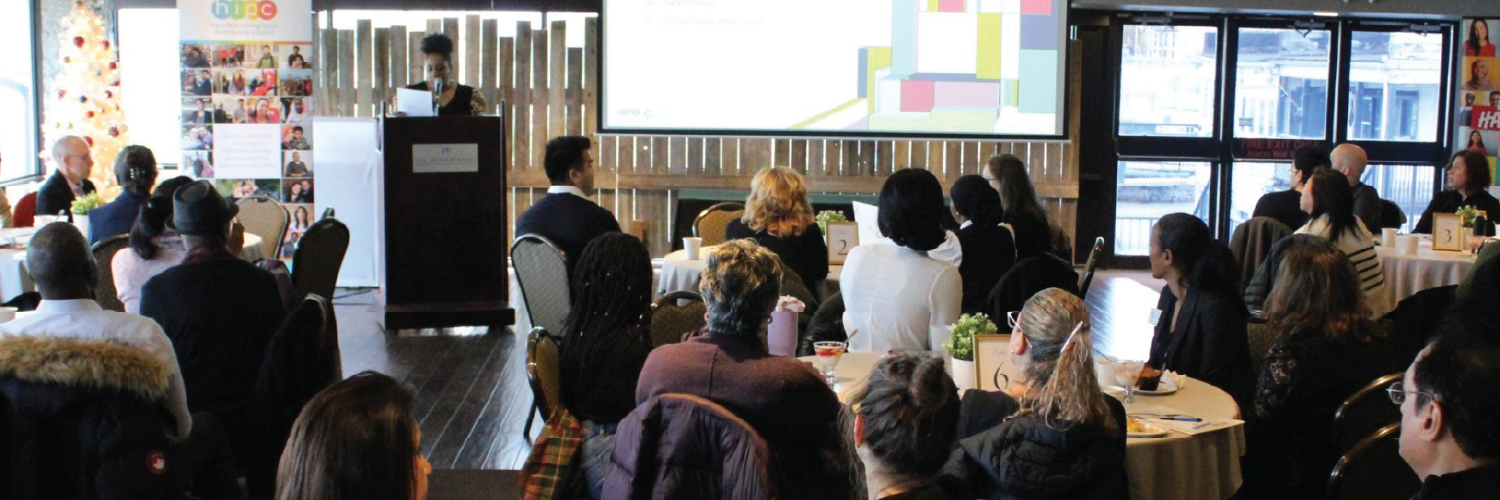 Photo from the 2025 community forum showing an audience facing away from the camera and a woman at a podium explaining a slide displayed on a screen.